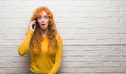 Young redhead woman standing over brick wall talking on the phone scared in shock with a surprise...
