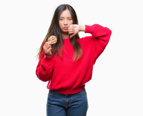 Young asian woman eating chocolate chip cookie over isolated background with angry face, negative sign showing dislike with thumbs down, rejection concept
