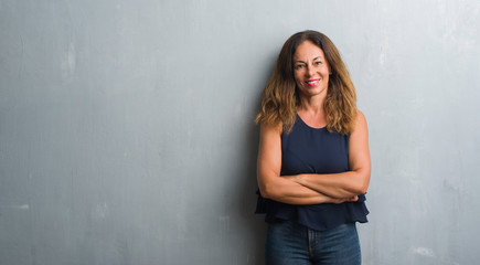 Middle age hispanic woman standing over grey grunge wall happy face smiling with crossed arms...