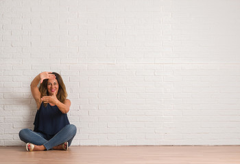 Middle age hispanic woman sitting on the floor over white brick wall smiling making frame with hands and fingers with happy face. Creativity and photography concept.