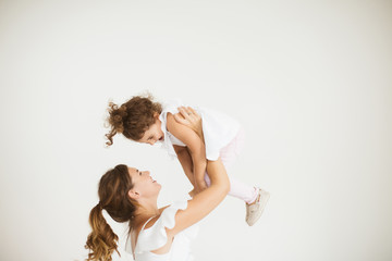 Young beautiful mother happily lifting little cute daughter up while spending time together over white background