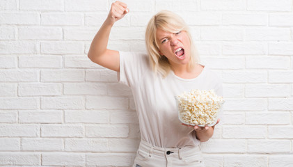 Adult caucasian woman over white brick wall eating pop corn annoyed and frustrated shouting with anger, crazy and yelling with raised hand, anger concept