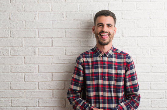 Young adult man standing over white brick wall sticking tongue out happy with funny expression. Emotion concept.