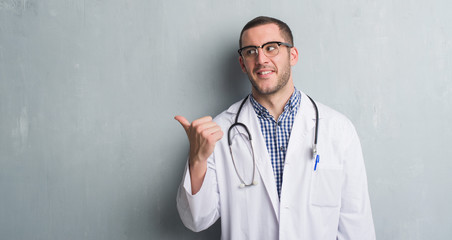 Young caucasian man over grey grunge wall wearing doctor uniform pointing and showing with thumb up to the side with happy face smiling