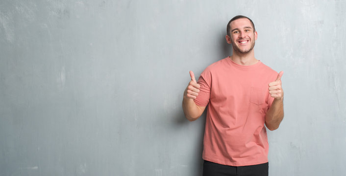 Young caucasian man over grey grunge wall success sign doing positive gesture with hand, thumbs up smiling and happy. Looking at the camera with cheerful expression, winner gesture.