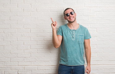 Young caucasian man standing over white brick wall wearing sunglasses very happy pointing with hand and finger to the side