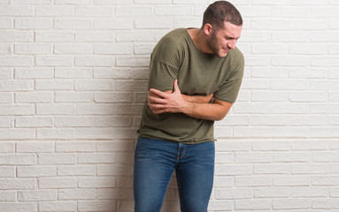Obraz na płótnie Canvas Young caucasian man standing over white brick wall with hand on stomach because nausea, painful disease feeling unwell. Ache concept.