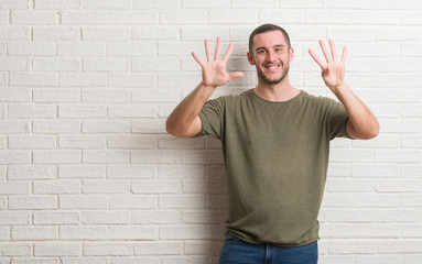 Young caucasian man standing over white brick wall showing and pointing up with fingers number nine...