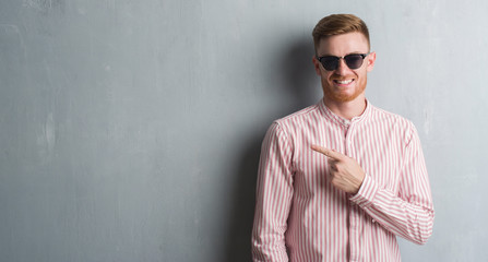 Young redhead man over grey grunge wall wearing sunglasses very happy pointing with hand and finger to the side