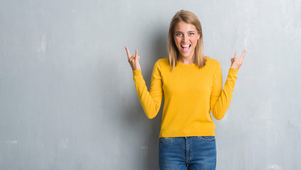 Beautiful young woman standing over grunge grey wall shouting with crazy expression doing rock symbol with hands up. Music star. Heavy concept.