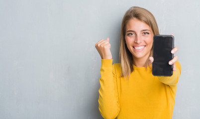 Fototapeta premium Beautiful young woman over grunge grey wall showing smartphone screen screaming proud and celebrating victory and success very excited, cheering emotion