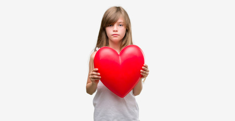Fototapeta na wymiar Young blonde toddler holding a red heart with a confident expression on smart face thinking serious