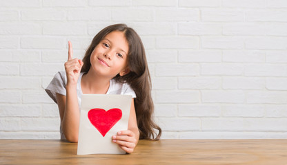 Young hispanic kid sitting on the table giving mother day card surprised with an idea or question pointing finger with happy face, number one