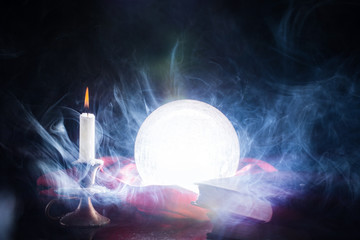 Magic crystal lights ball on table with candle in candlestick and books, ball is smokes and on black background. Halloween concept. Close up, selective focus