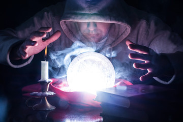 Wizard with hood and lights smoke magic crystal ball on desk with candle in candlestick and old books predicting the future by looking into ball and holding hand above tools. Close up, selective focus