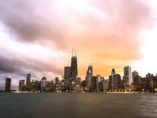 A beautiful dramatic sunset photograph of downtown Chicago over Oak Street beach with smooth waters of Lake Michigan in the foreground and colorful orange and pink sky above.