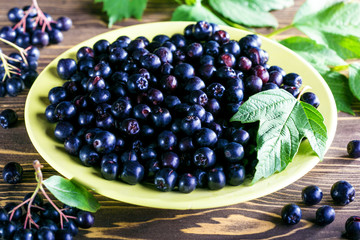 Aronia melanocarpa or black chokeberry with leaves in yellow bowl on wooden background. Healthy natural  food.