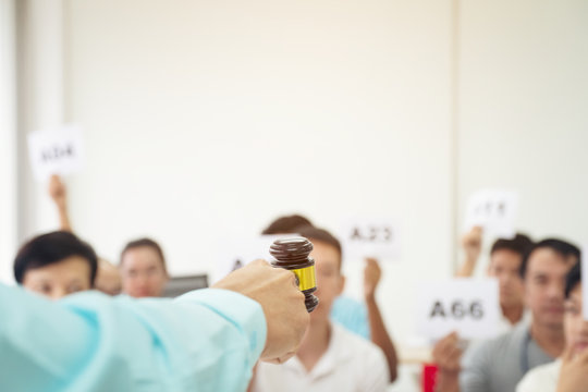 Close up auctioneer hand, holding gavel, wooden hammer, and blur group of people in auction room, one man raising hand up for bidding. Product or project auction market concept background