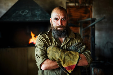 Portrait of a brutal, confident blacksmith, a man with a beard. Standing in the workshop, folding...