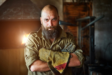 Portrait of a brutal, confident blacksmith, a man with a beard. Standing in the workshop, folding his arms over his chest.