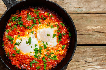 Homemade breakfast shakshuka - fried eggs, onion, bell pepper, tomatoes and parsley in cast-iron frying pan on rustic wooden table. Jewish cuisine. Top view