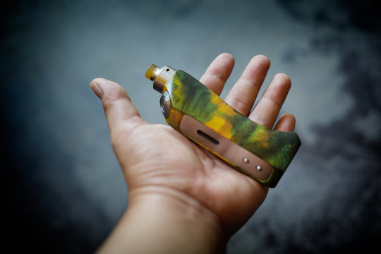 high end yellow green stabilized wood box mods with rebuildable dripping atomizer in hand on dark grey texture background, vaping device, vape gear, vaporizer equipment, selective focus