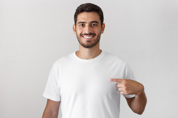 Handsome young man pointing with index finger at blank white t-shirt with empty space for your advertising text or image, standing isolated on gray background