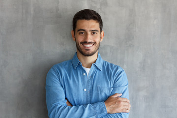 Smiling man in blue shirt, standing with crossed arms against gray textured wall