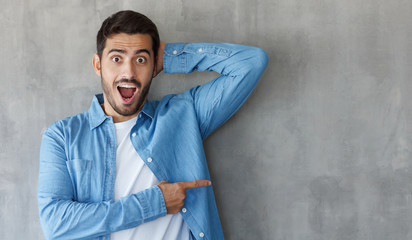 Horizontal photo of young caucasian man isolated on gray background dressed in blue shirt looking at camera with mouth and eyes open with surprise, pointing right, copyspace for ads