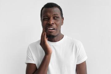 Young african american man in white t shirt, isolated on gray background with toothache, touching his face with expression of horrible suffer from health problem and aching tooth