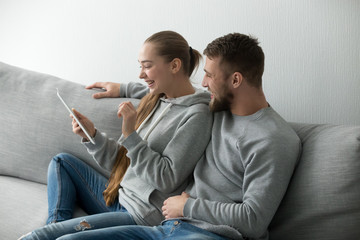 Smiling millennial couple sitting on sofa hugging watching funny video on tablet, young boyfriend...
