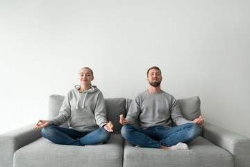 Happy millennial couple sit in lotus position on home coach, calm boyfriend and girlfriend meditate practicing yoga on sofa, mindful man and woman relieve stress breathing fresh air with mudra hands