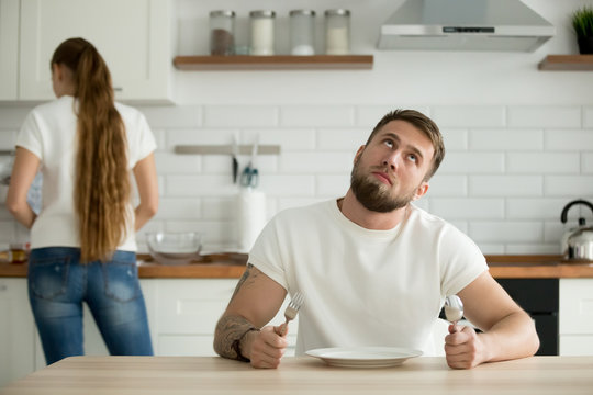 Lazy millennial husband look bored waiting for dinner or breakfast cooked by wife, indifferent man wanting eat sitting with empty plate at table thinking about something rolling eyes looking bored