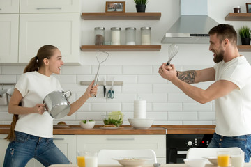 Happy young couple having fun in kitchen playing with kitchenware while preparing breakfast,...