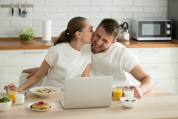 Loving young girlfriend kiss millennial boyfriend while having tasty healthy oatmeal porridge for breakfast, romantic couple cuddle enjoying delicious food in the morning at home watching laptop