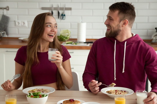 Happy young couple smiling enjoying delicious healthy oatmeal breakfast in kitchen, excited boyfriend and girlfriend eating tasty porridge, having homemade food, having fun in the morning at home