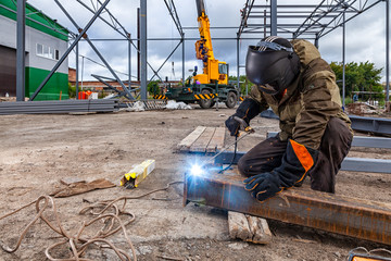 Man welder in welding mask, building uniform and blue protective gloves brews metal welding machine...