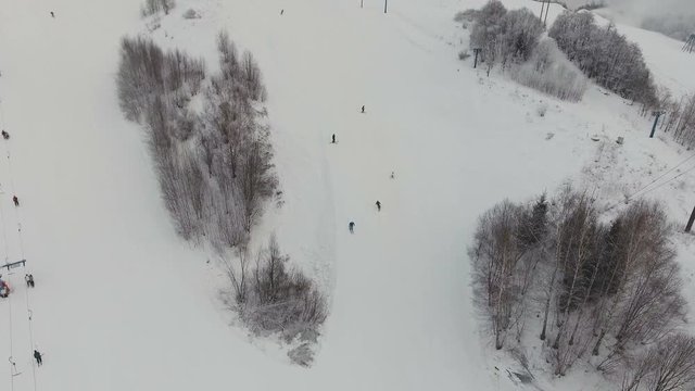 Aerial view: Skiers and snowboarders going down the slope in winter day. Skiers and snowboarders enjoying on slopes of ski resort in winter season. 4K video, aerial footage.