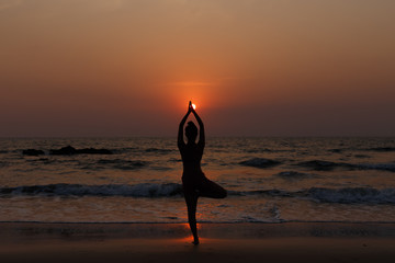 Fototapeta na wymiar Young slender girl with her back doing yoga at sunset on the beach. Pose on balance, pose tree. Hands are raised and connected together. Natural backlight.