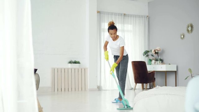 Pretty African American girl is mopping floor at home with flat plastic mop wearing gloves and wear-at-home clothing. Modern interiors, cleanness and people concept.