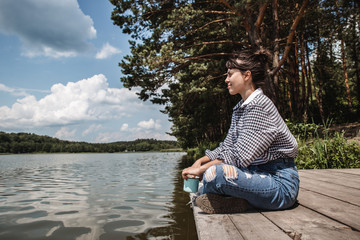 Fototapeta premium young adult woman sitting on wooden dock drinking coffee and looking at lake