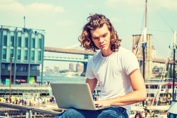 Young Hispanic American Man traveling, working in New York, with long curly hair, wearing white T shirt, sitting by East River, working on laptop computer, thinking. Bridges, boats on background..
