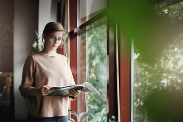 Indoor shot of good-looking serious european female coworker in trendy and cozy pullover, holding magazine and gazing with dreamy expression at camera, standing near window, dreaming and relaxing