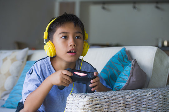 Young Latin Little Child Excited And Happy Playing Video Game Online With Headphones Holding Controller Having Fun Sitting On Couch In Kid Gaming Addiction