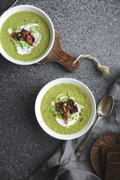 Overhead image of creamy green pea soup with fried bacon and herbs on gray background
