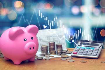 Coins stack on saving account book from bank with pink piggy bank and calculator on wooden table with candlestick chart suitable for finance and investment concept and blue bokeh background.