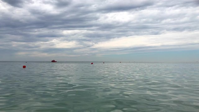 Red buoy float and red lifeboat remain afloat on calm aquamarine sea water  in the distance under low grey clouds on sky, hole in clouds
