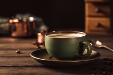 Green cup of coffee with beans and coffee mill on old wooden background with copy space. Retro style toned dark photography.