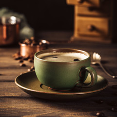 Green cup of coffee with beans and coffee mill on old wooden background with copy space. Retro style toned dark photography.