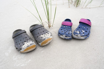 Two sand covered colorful rubber clog shoes on white beach sand with green blades of grass -...